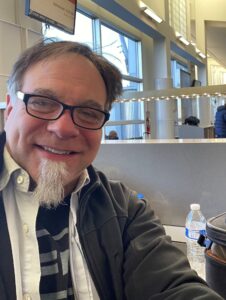 A man with glasses and a goatee smiles at the camera in a modern waiting area, with a water bottle and bag on the counter beside him.