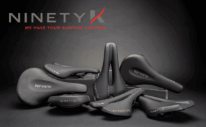 A variety of black NINETYK bicycle saddles are displayed on a dark background with the NinetyK logo and slogan, "WE HAVE YOUR SURFACE COVERED," visible above.
