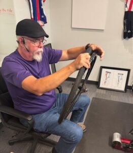 A man sitting in an office chair works on a bicycle wheel, wearing a headset and purple shirt, with cycling clothes and framed sketches in the background.